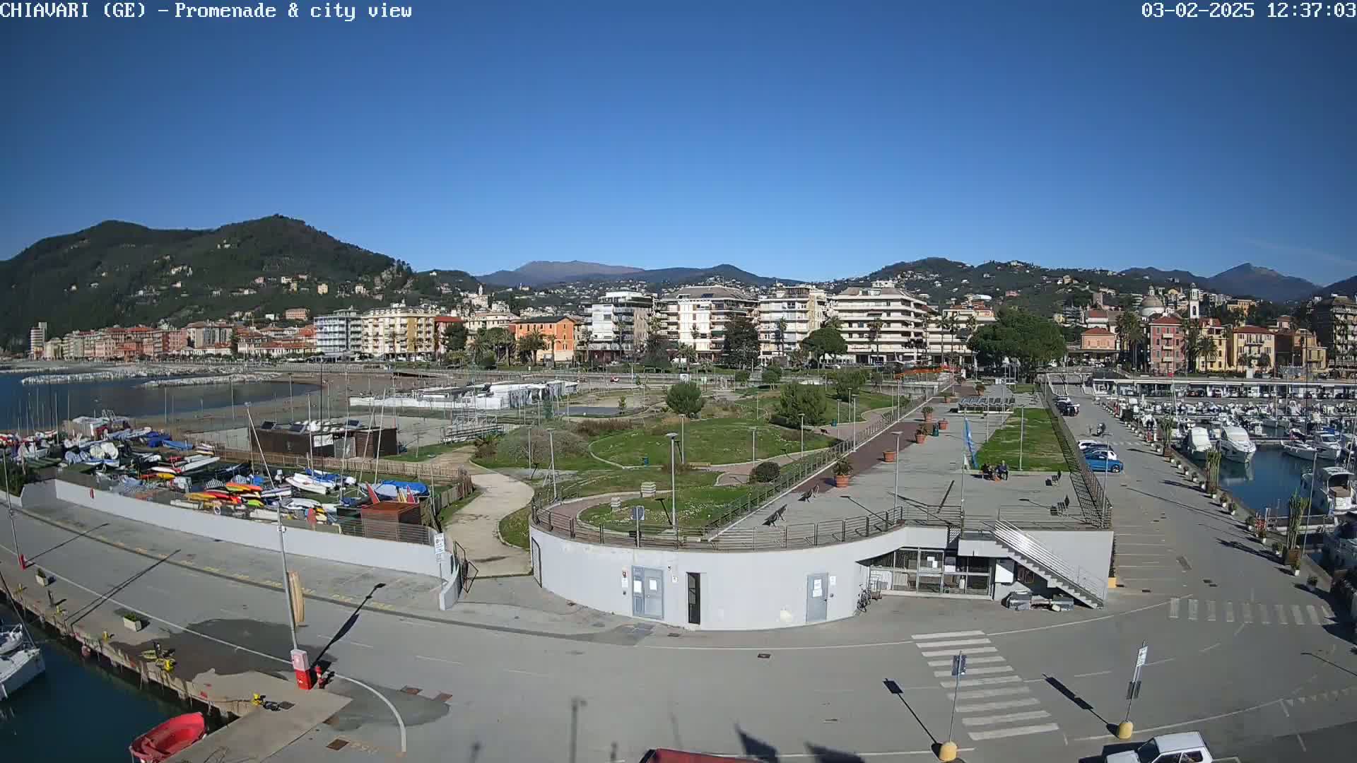 A waterfront promenade with a marina, park, and residential buildings in the background under a clear blue sky.