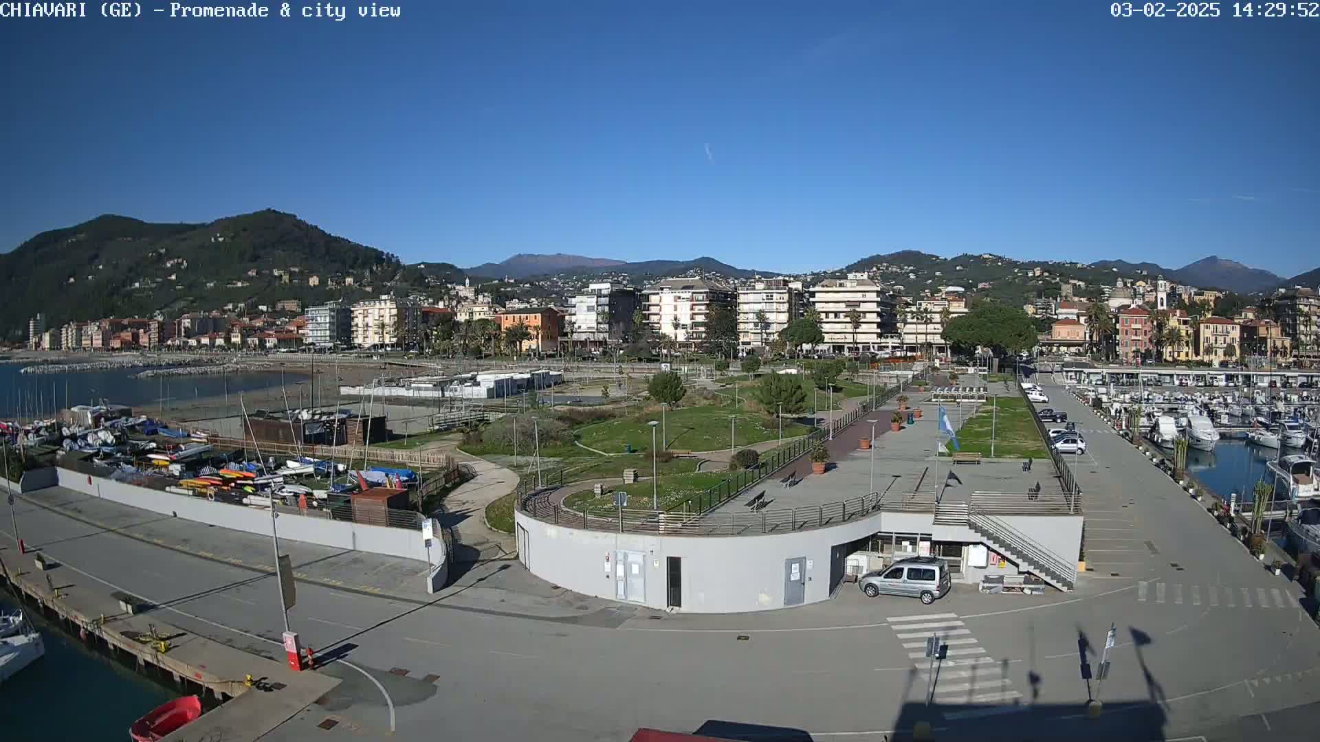 A coastal town with a marina, park, and buildings stretches along a bay under a clear blue sky.
