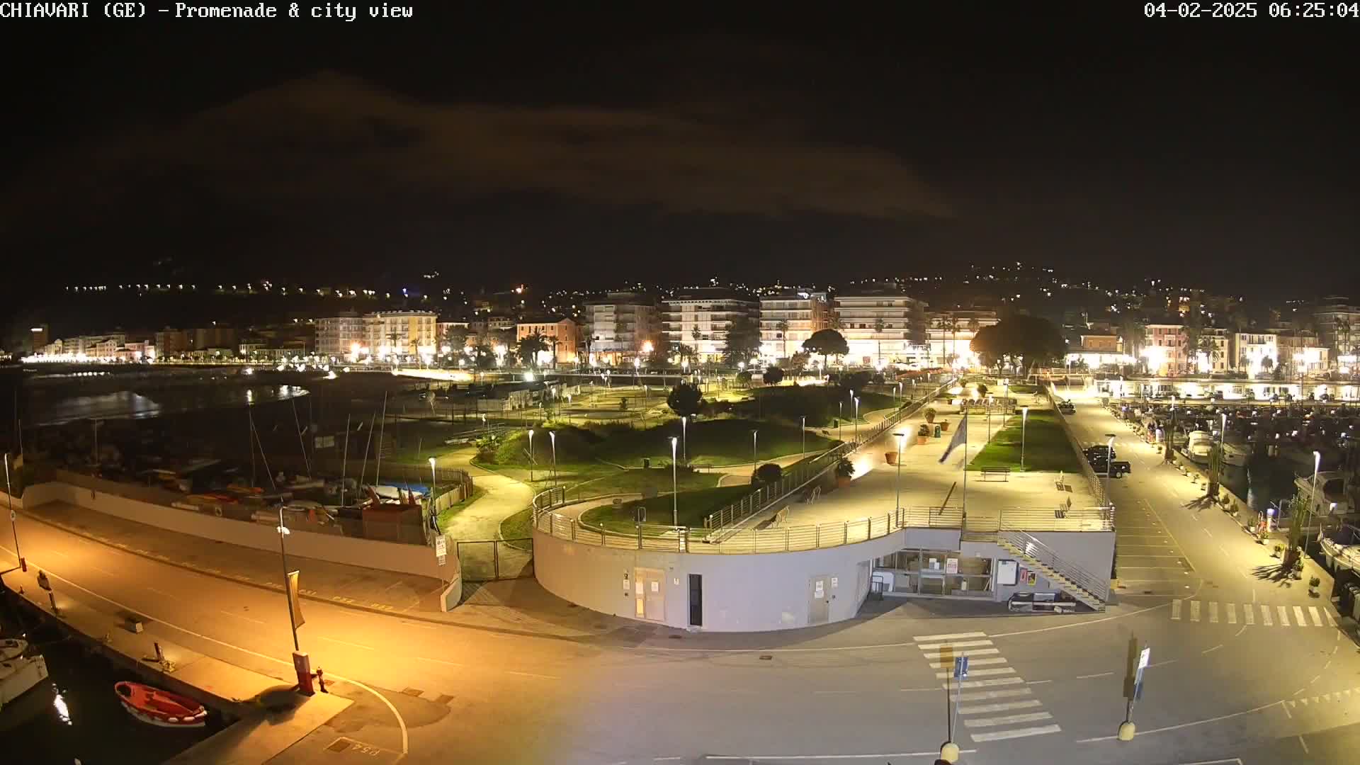 A nighttime aerial view shows a brightly lit city waterfront with a promenade, park, and marina.