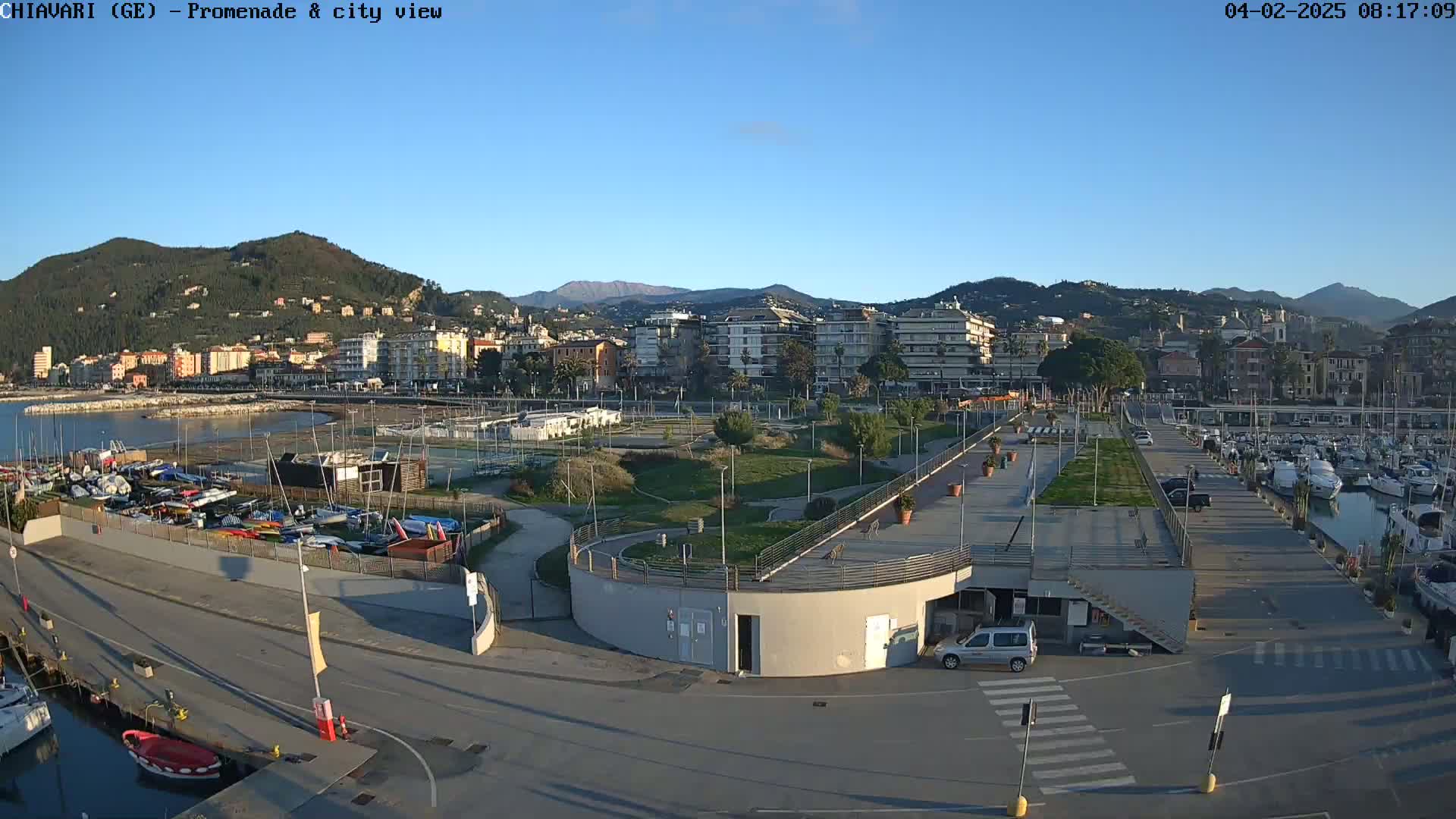 A seaside town with a harbor full of boats, a promenade, and a park under a clear blue sky.