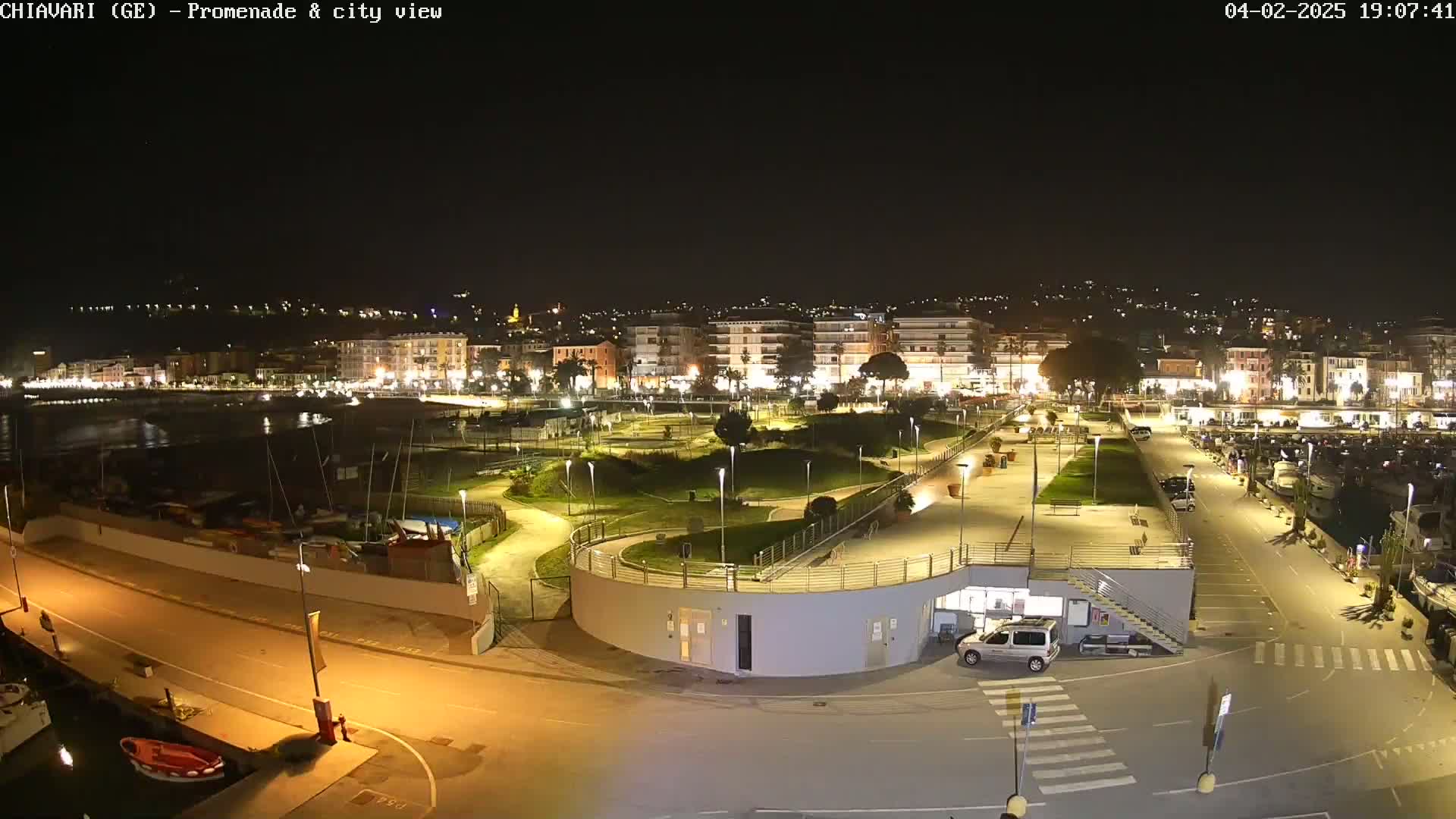 A nighttime view of a brightly lit waterfront promenade and city, with a marina filled with boats in the background.