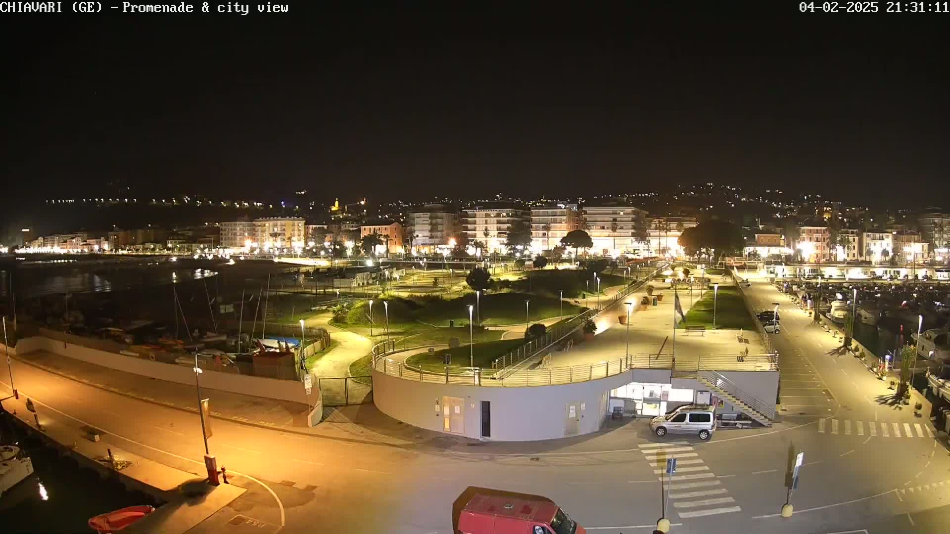 A nighttime, aerial view of a city's waterfront promenade, featuring a marina with boats, a park with winding paths, and buildings lit up along the shoreline.