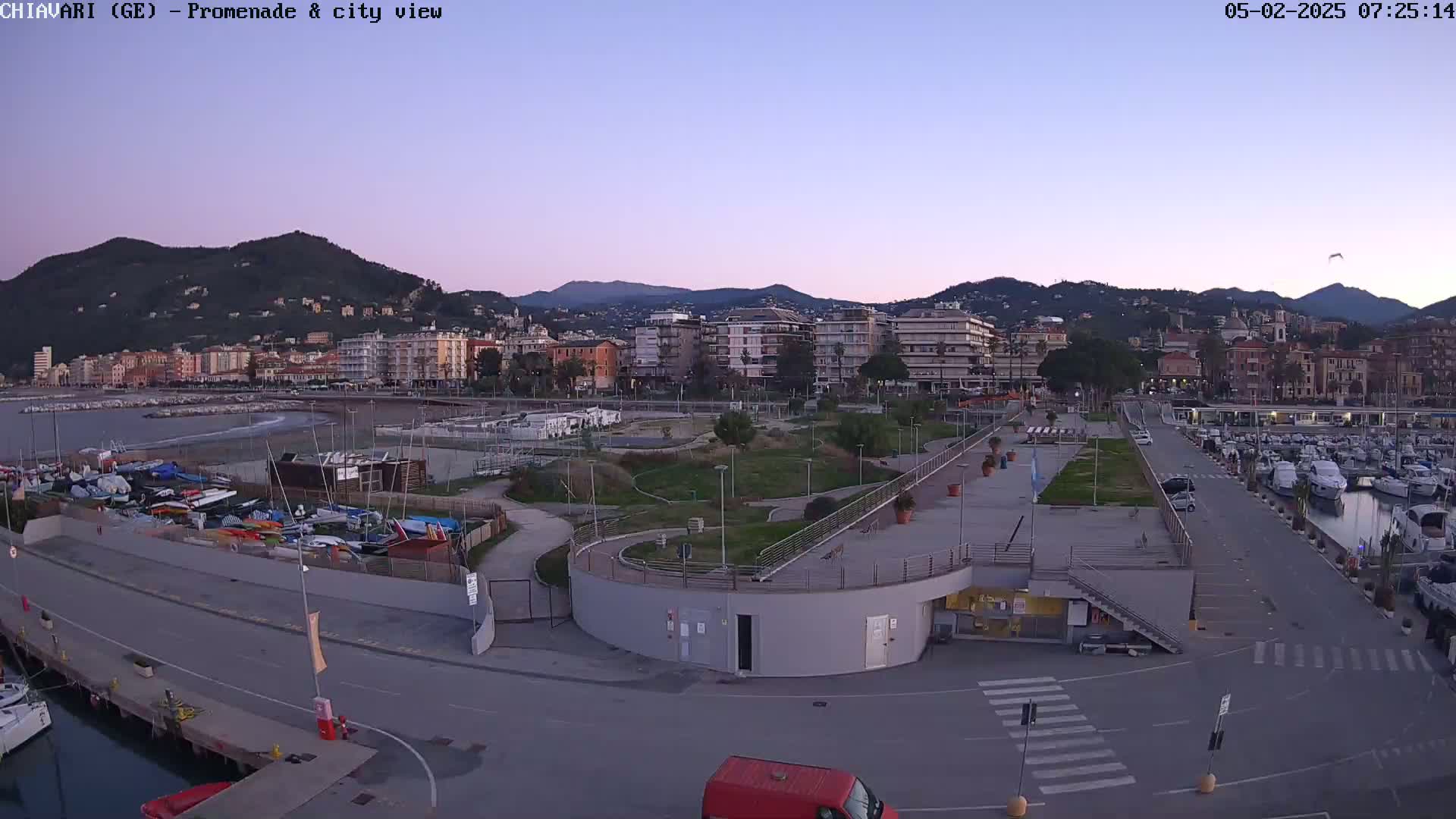 A coastal city at dawn, with calm waters, a marina filled with boats, and a promenade winding through a park-like area next to a waterfront lined with buildings.
