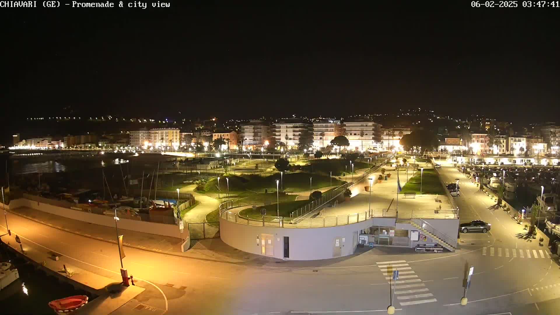 A nighttime, brightly lit waterfront promenade and city view shows a harbor with boats, a park with winding paths, and buildings along the shoreline.