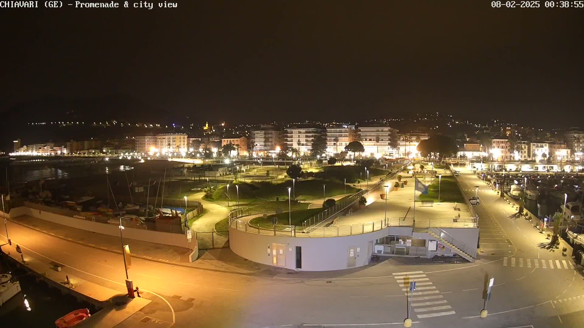 A nighttime, brightly lit view of a harbor promenade and city skyline overlooking a calm body of water.