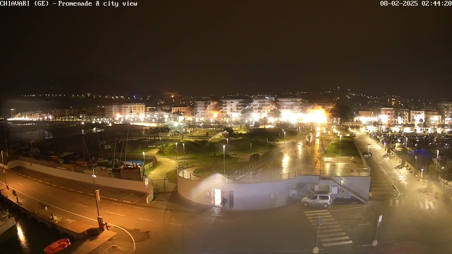 A nighttime, rain-slicked coastal town scene shows a promenade, park, parking lot, and buildings illuminated by streetlights, with a dark sea visible in the background.