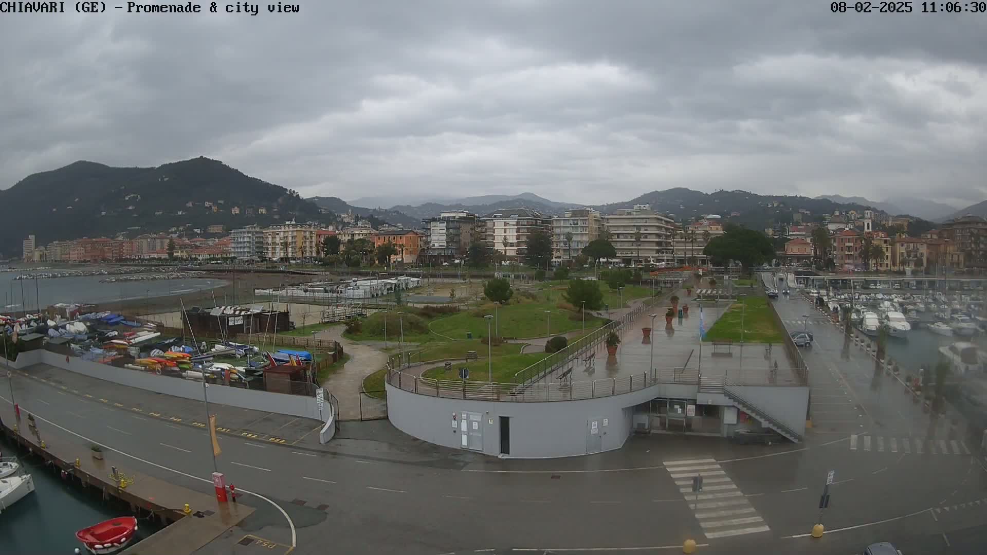 A rain-slicked waterfront promenade overlooks a marina filled with boats and a city nestled against a backdrop of mountains under a cloudy sky.