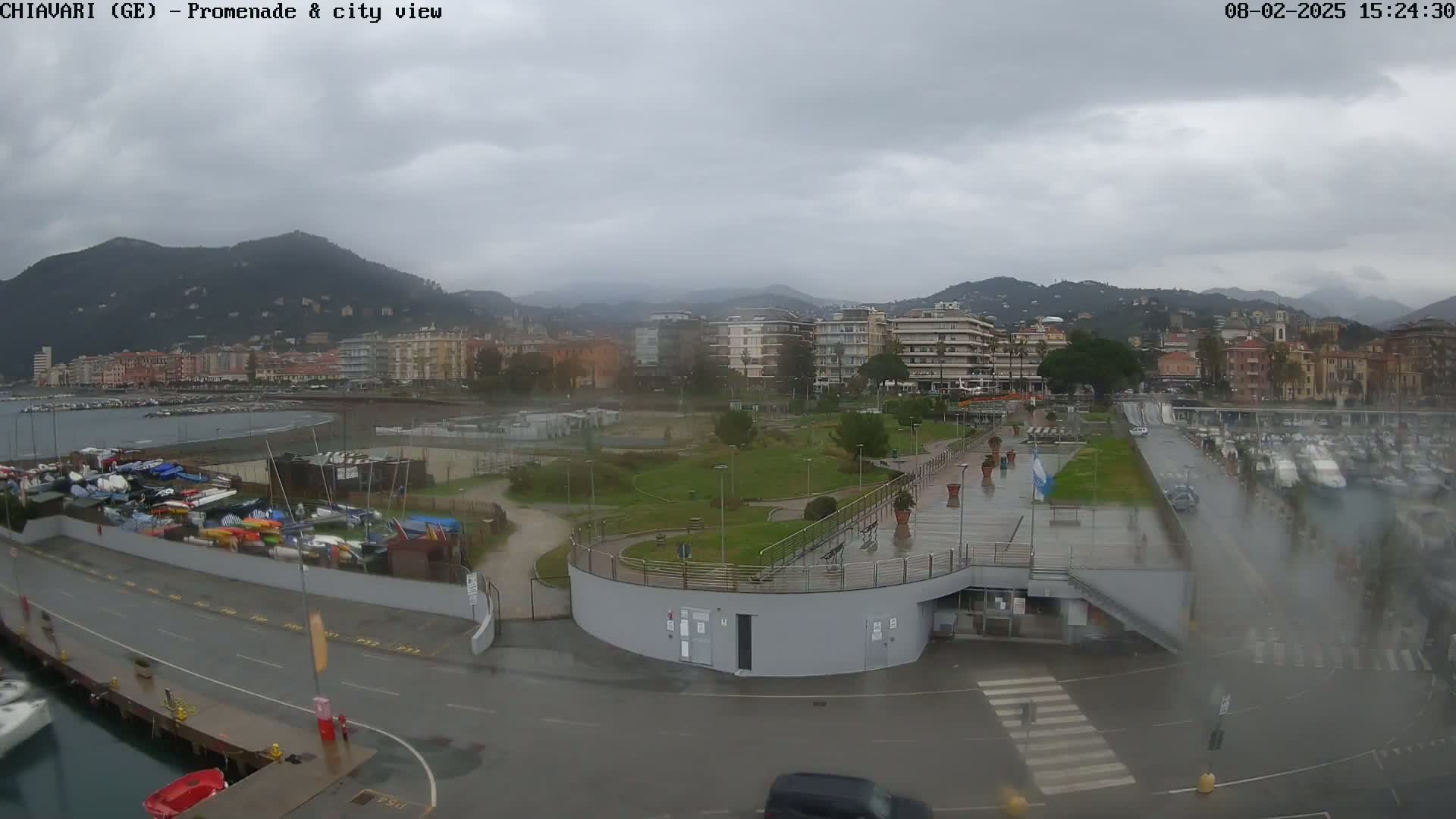 A rain-slicked waterfront promenade overlooks a city nestled against a mountain range under a cloudy sky.
