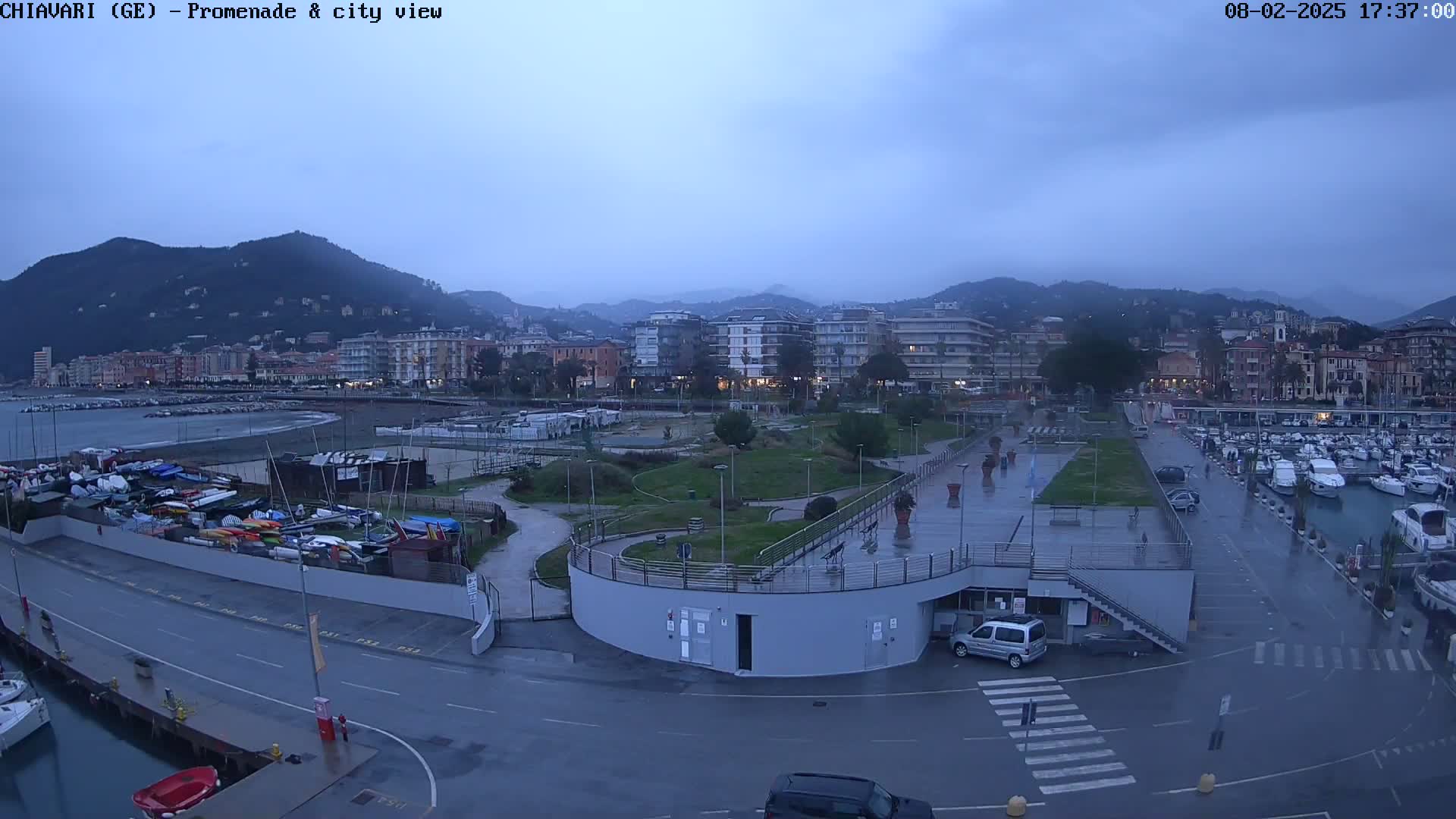 A wet city promenade and marina are shown under an overcast sky, with boats moored in the harbor and buildings along the waterfront, backed by hills.