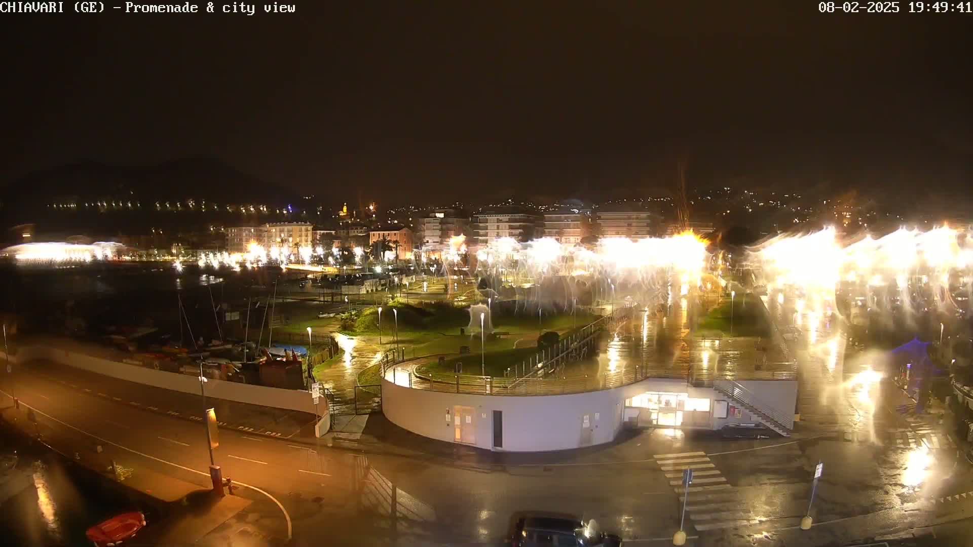 A nighttime view of a waterfront city promenade with a water feature displaying bright lights in the rain.