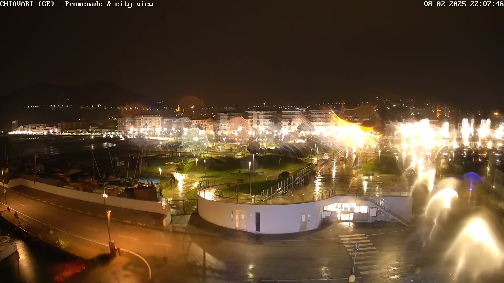 A nighttime view of a rain-slicked seaside promenade and city, featuring illuminated buildings and numerous water features.