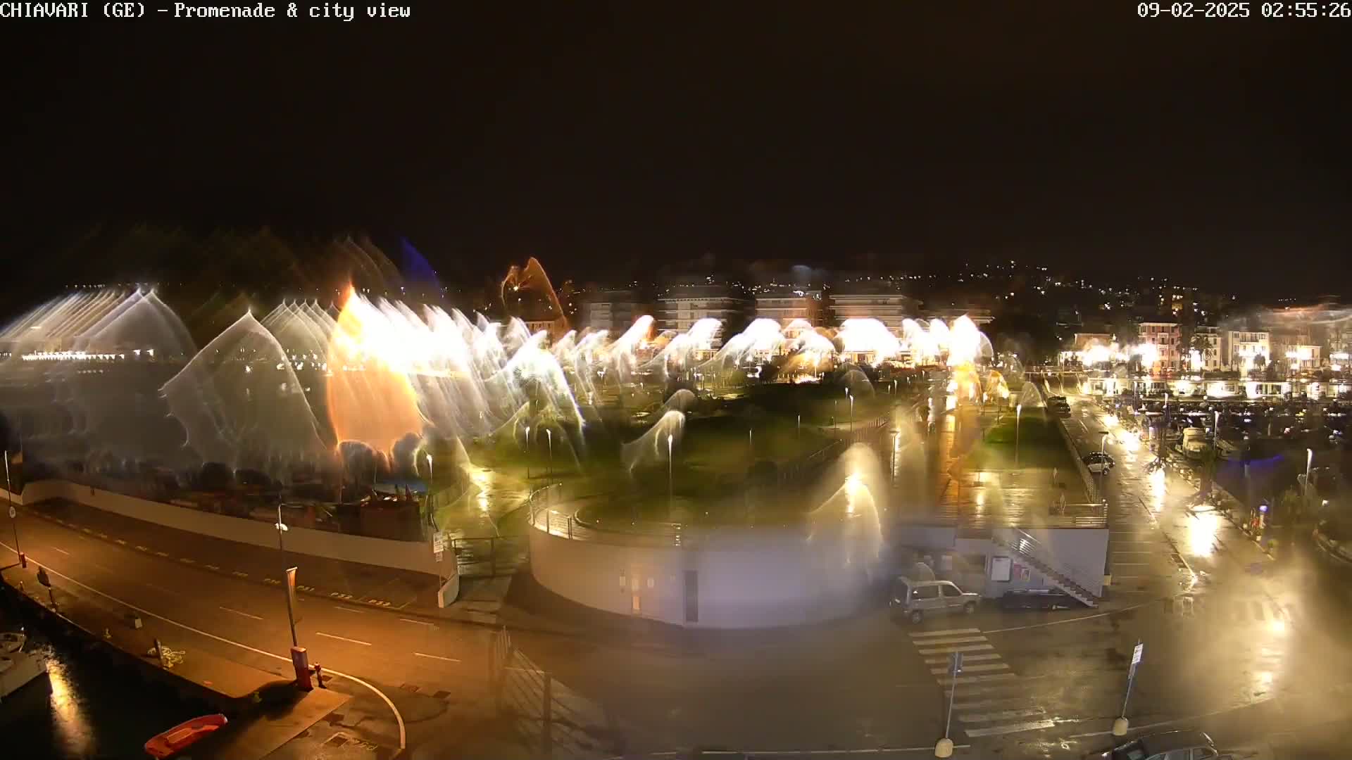 A nighttime view of a city promenade with a large water fountain display illuminated with various colors under wet conditions.