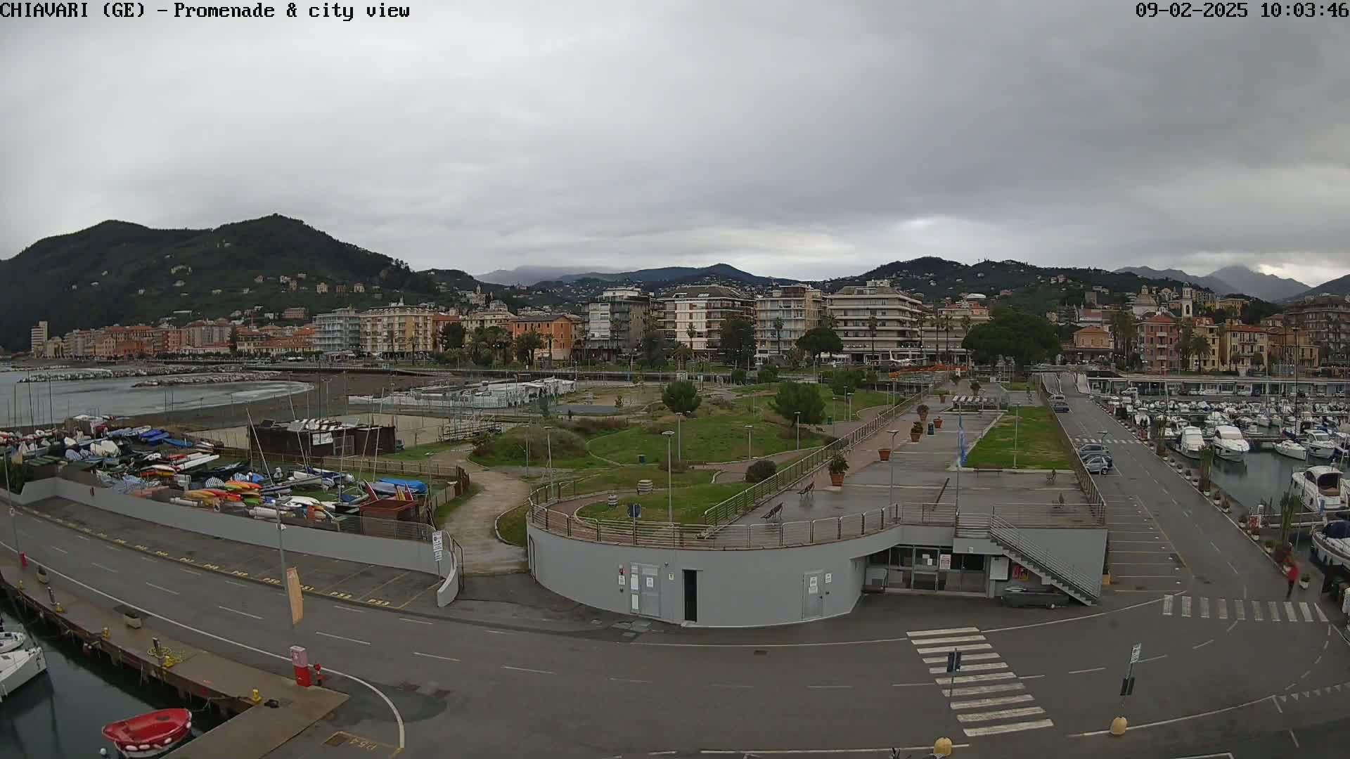 An overcast day reveals a coastal city scene with a marina filled with boats, a promenade, a park-like area, and buildings extending into the background against a backdrop of mountains.