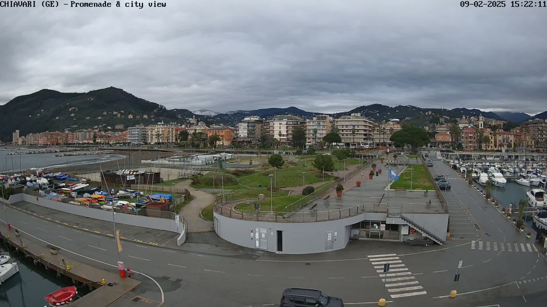 An overcast day reveals a harbor filled with boats, a promenade with green spaces, and a city nestled against mountains in the background.