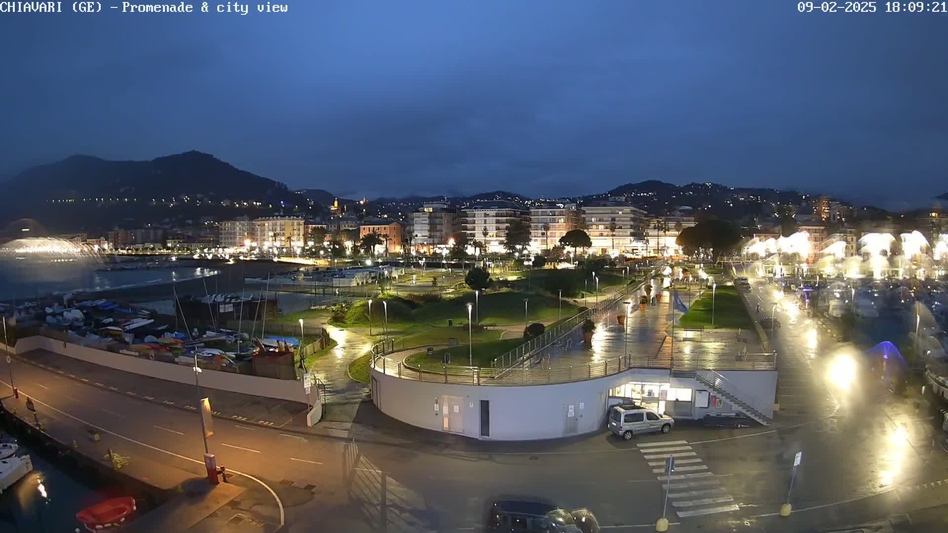 A nighttime view of a waterfront city illuminated by lights, with a wet promenade and a water fountain in the distance under a dark, cloudy sky.