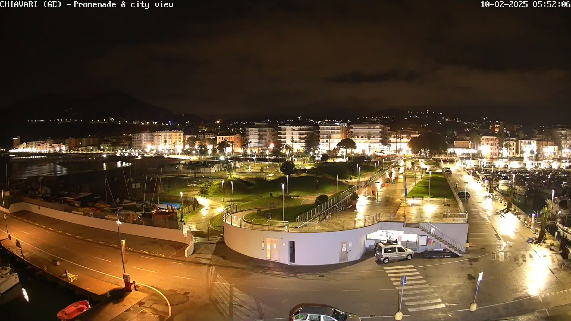 A nighttime view of a city's waterfront promenade and harbor, illuminated with lights, showing wet pavement under a dark sky.