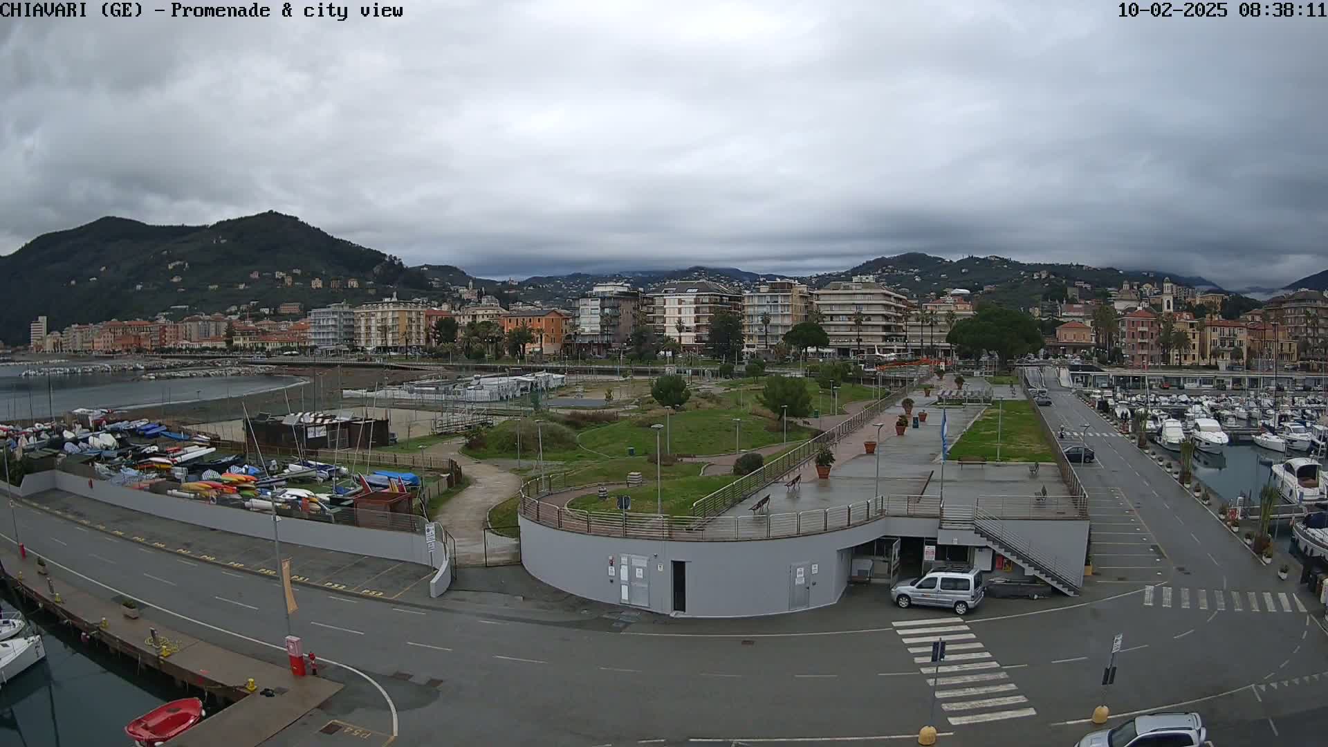 An overcast day reveals a harbor filled with boats, a promenade, and a town nestled against a mountain range.