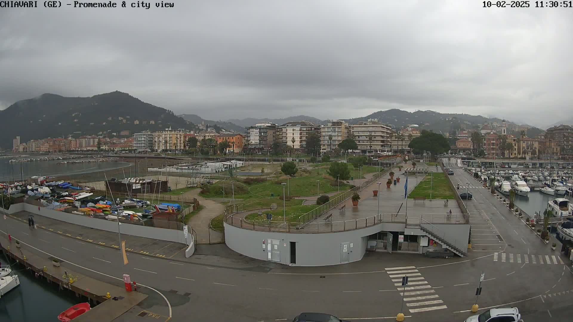 A waterfront promenade overlooks a harbor filled with boats under a cloudy sky, with a city and hills in the background.