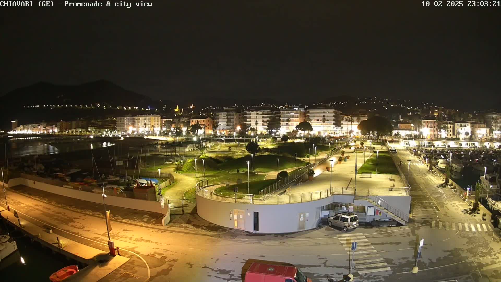 A nighttime, aerial view of a city's waterfront promenade and marina, featuring a park, buildings, and boats, under clear skies.