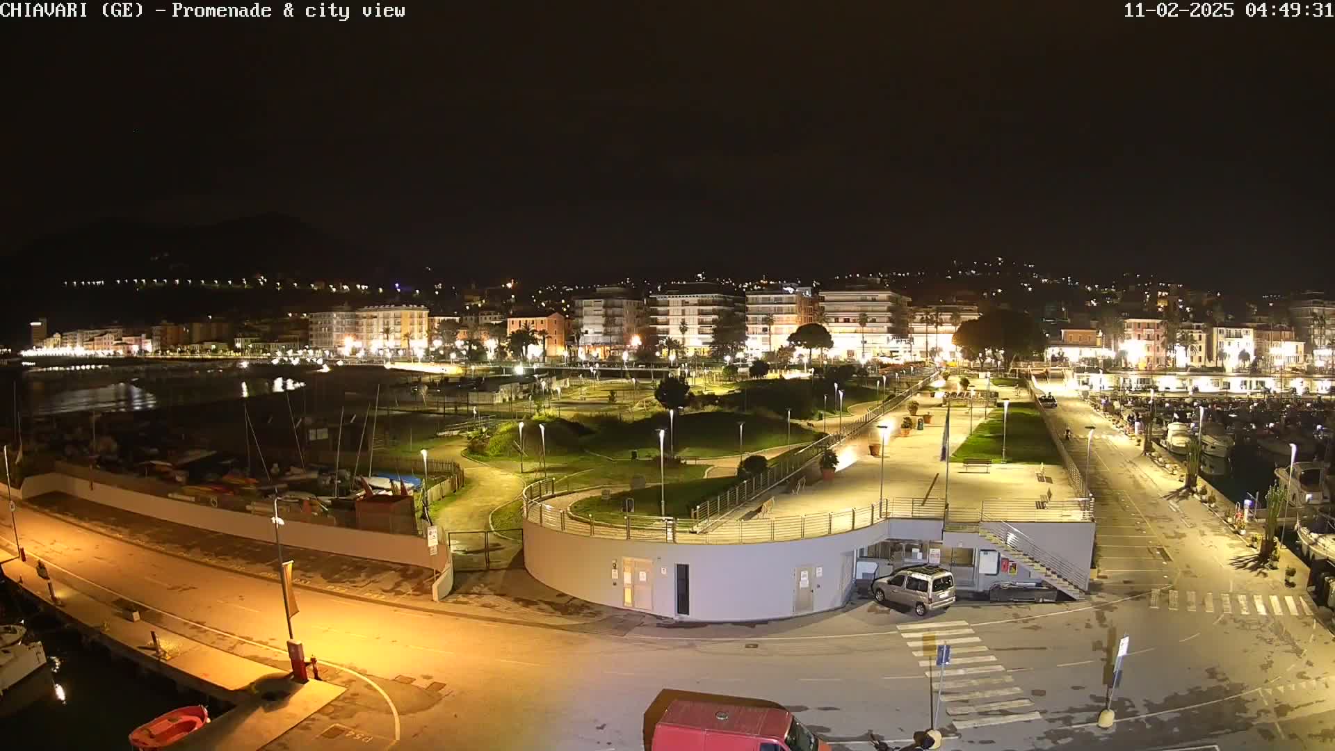 A nighttime aerial view of a city's waterfront promenade and harbor, illuminated by streetlights, with boats moored in the harbor and buildings lining the shore.
