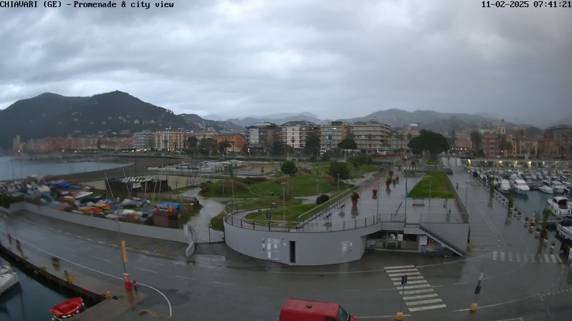 A wet city promenade and marina overlook a coastal town nestled between mountains under a cloudy, overcast sky.