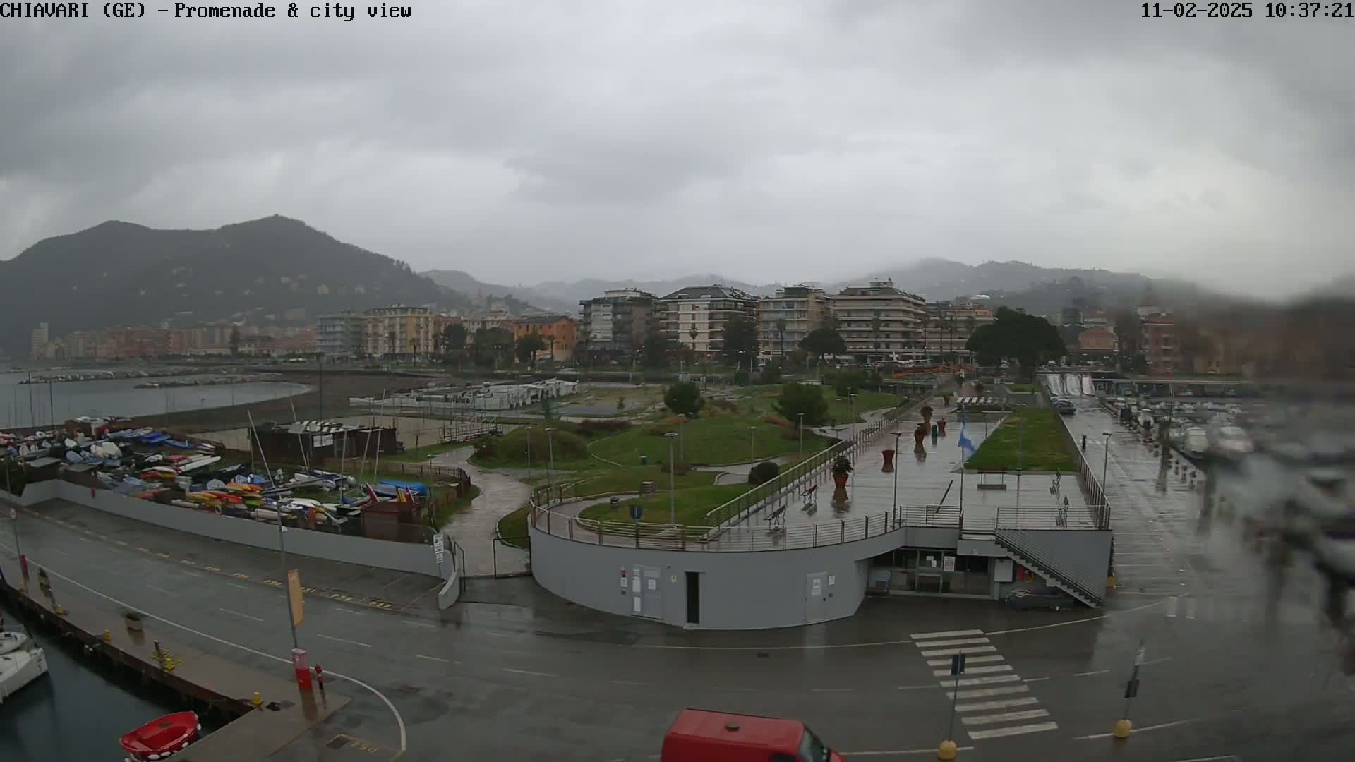 A rainy view of a waterfront promenade with a park, boat storage, buildings, and a parking lot, all under a gray, overcast sky with mountains in the background.
