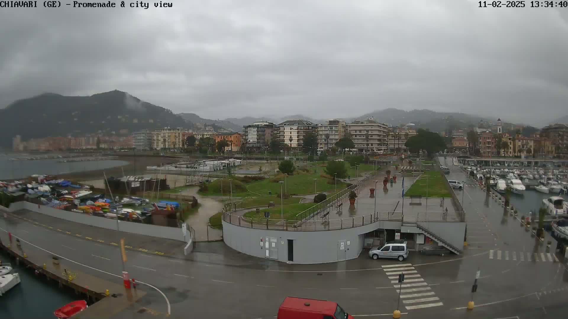A rainy view of a city waterfront, showing a promenade, park, marina with boats, and buildings set against a backdrop of hills.