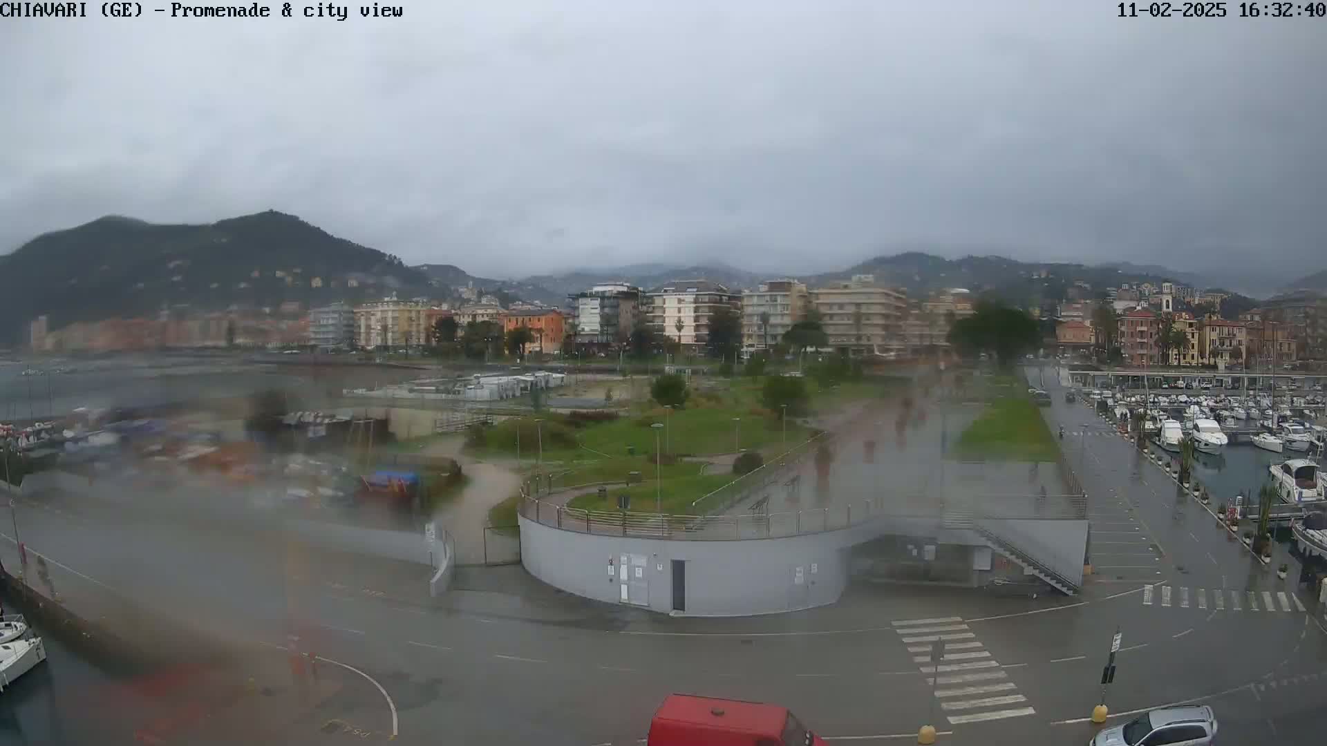 A rain-slicked waterfront promenade and marina overlook a town nestled against a mountain range under a cloudy sky.