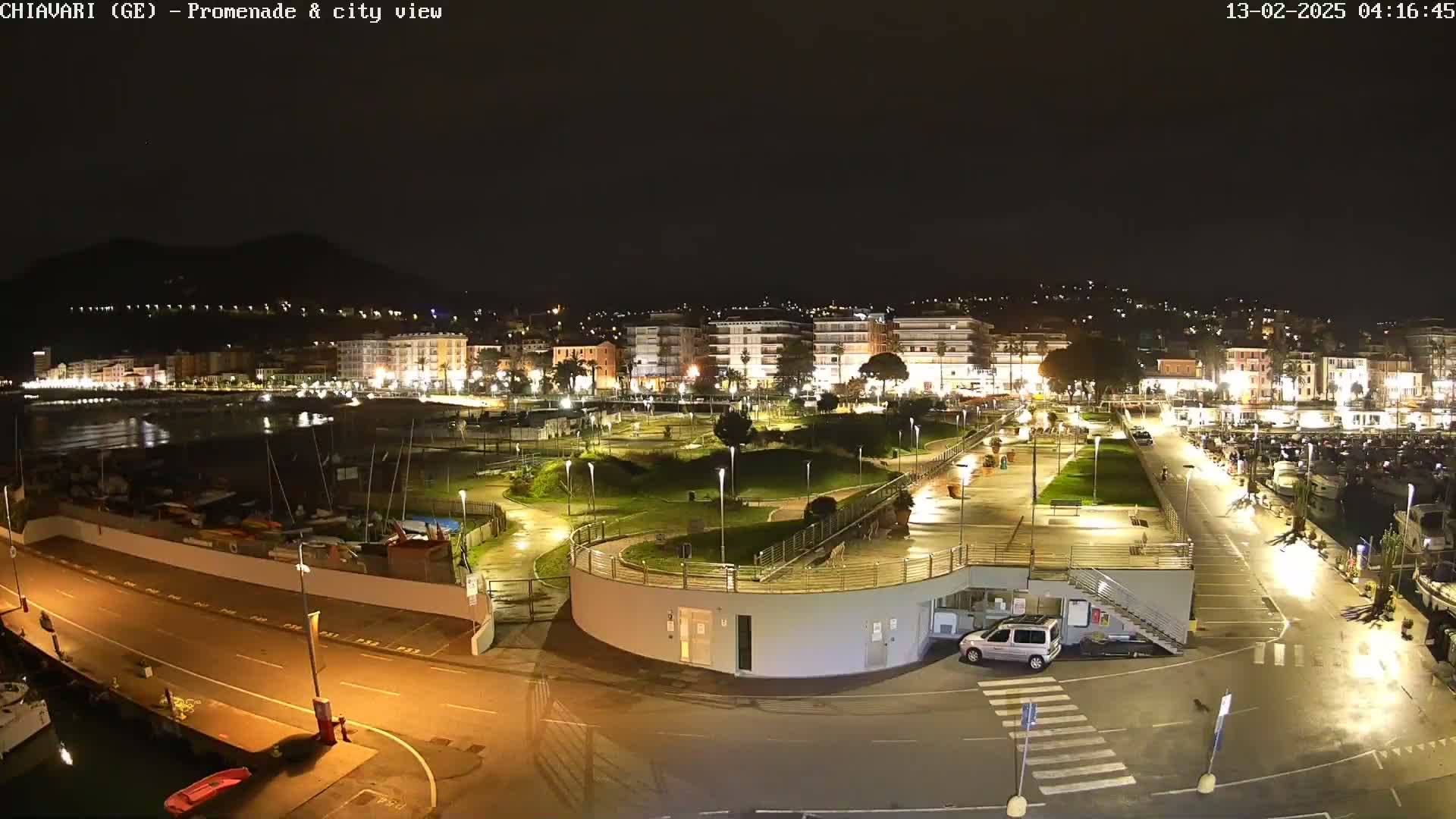 A nighttime view of a harbor town's waterfront promenade and marina, illuminated by streetlights, with a calm sea and boats docked.