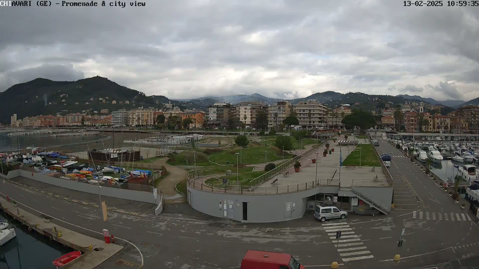 An overcast day shows a city's waterfront promenade with a marina, park, and buildings nestled against hills.