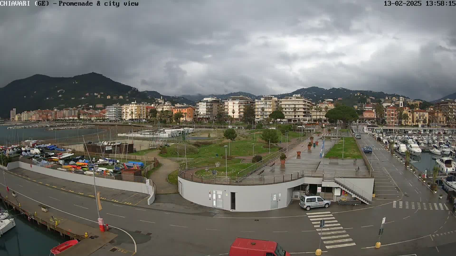 A coastal city is seen under an overcast sky, with a harbor filled with boats and a promenade adjacent to a park-like area.
