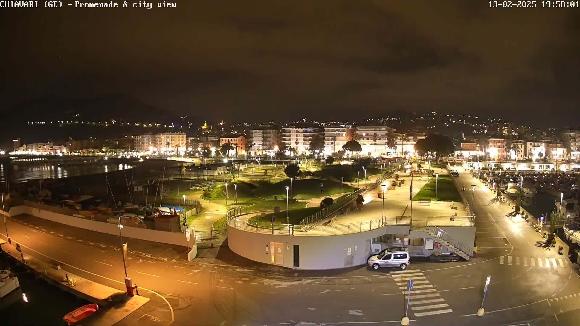 A nighttime view of a city's waterfront promenade and harbor, illuminated with streetlights, under a dark, cloudy sky.