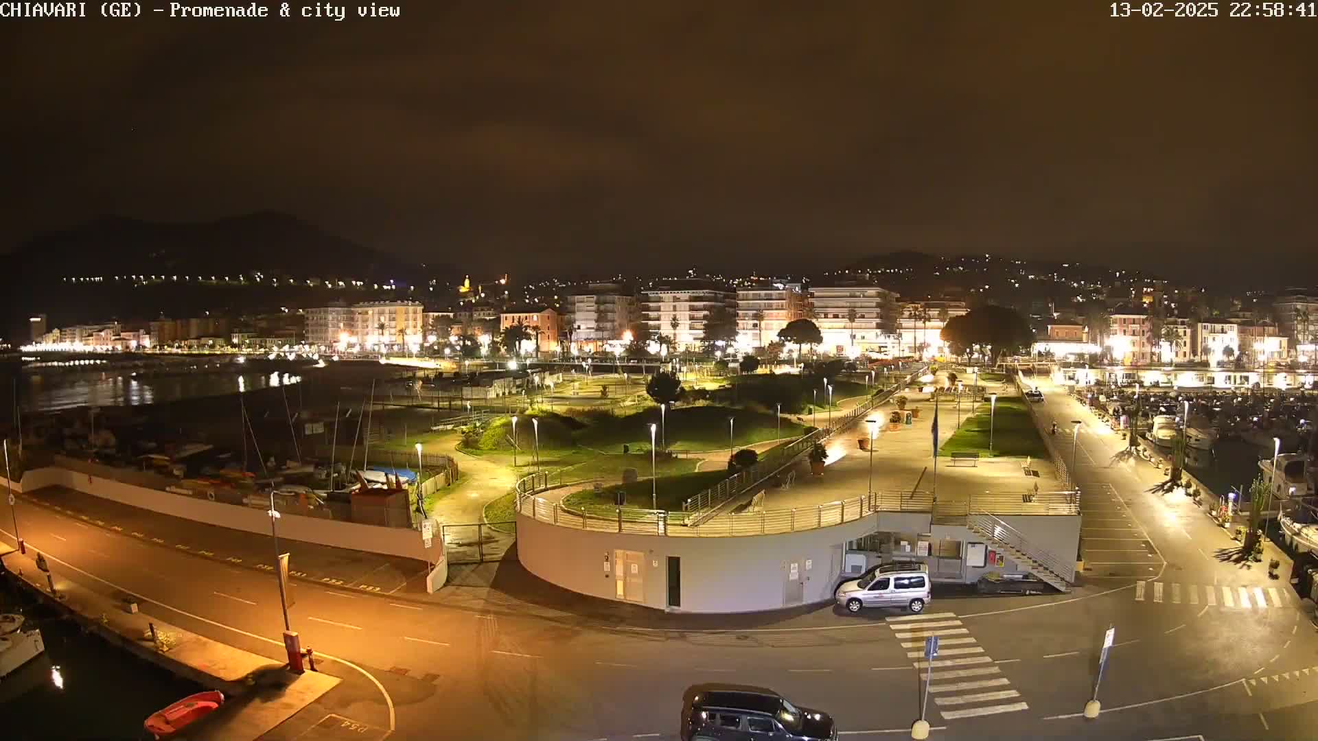 A nighttime aerial view of a city's illuminated waterfront promenade and harbor, featuring a landscaped park and parked cars.