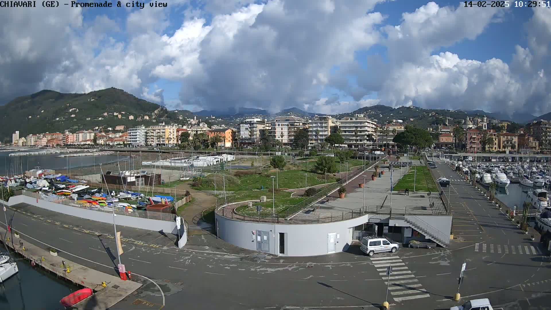 A harbor scene on a partly cloudy day shows a marina filled with boats, a promenade, and a town nestled against hills in the background.