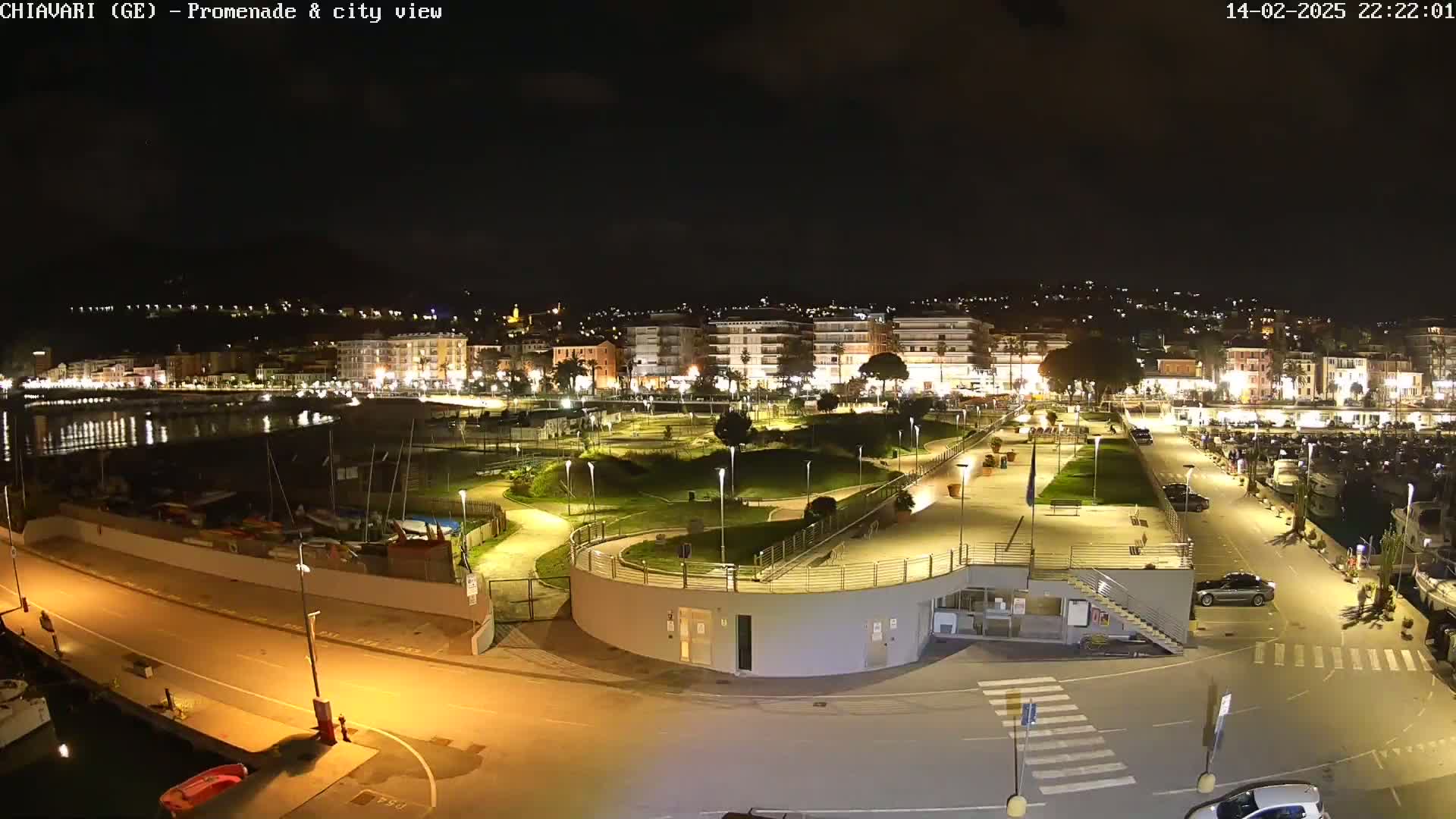A nighttime, high-angle view of a city's waterfront promenade and marina, featuring a park, buildings, and parked cars under clear skies.