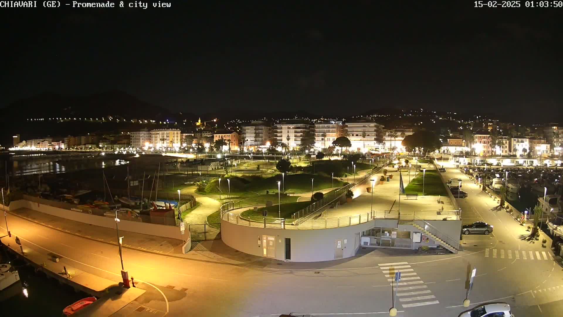 A nighttime aerial view shows a city's illuminated waterfront promenade, harbor, and park, with a dark sky.