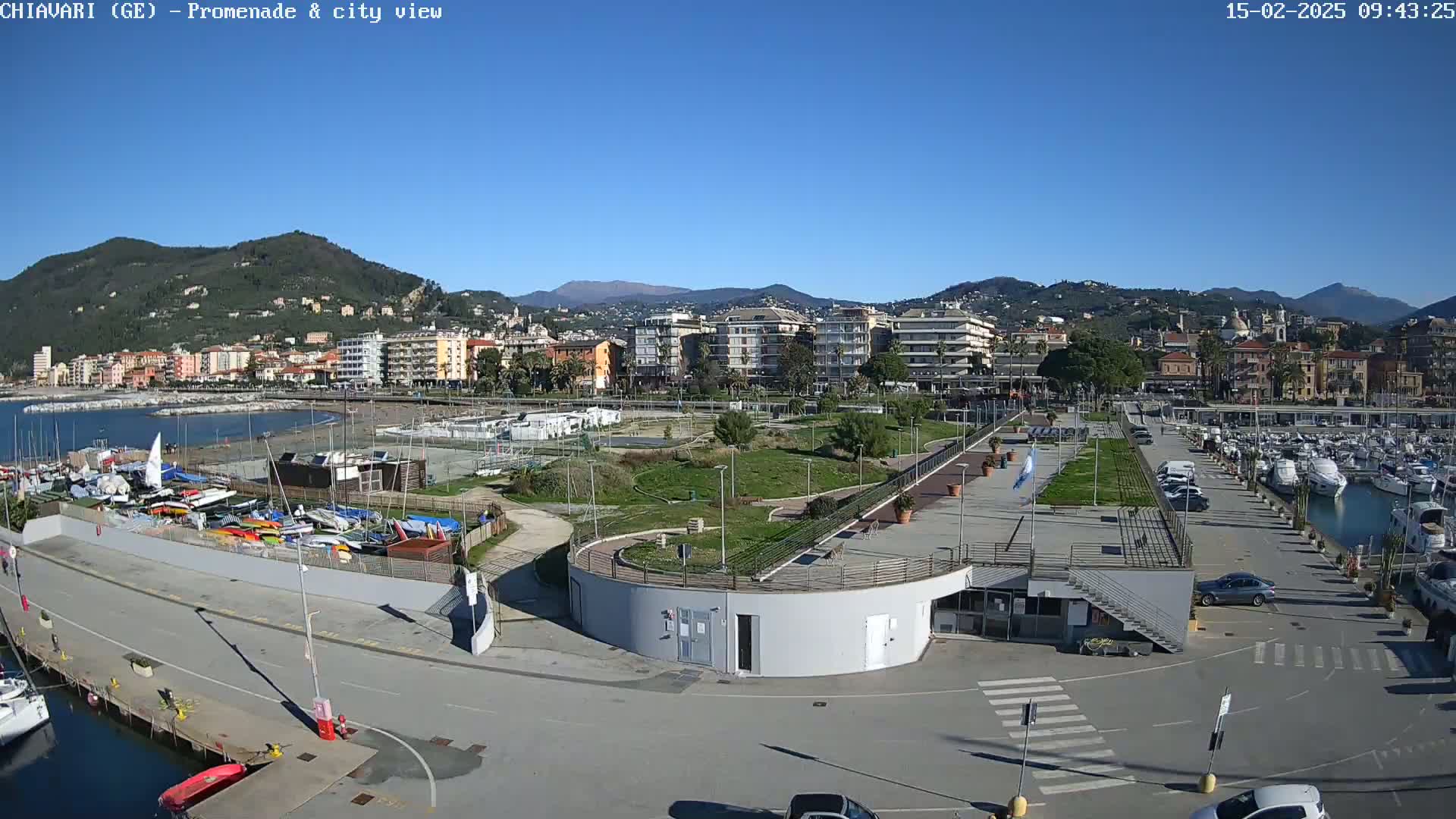 A seaside town under a clear blue sky, featuring a marina filled with boats, a promenade, a park, and buildings stretching along the coast backed by mountains.