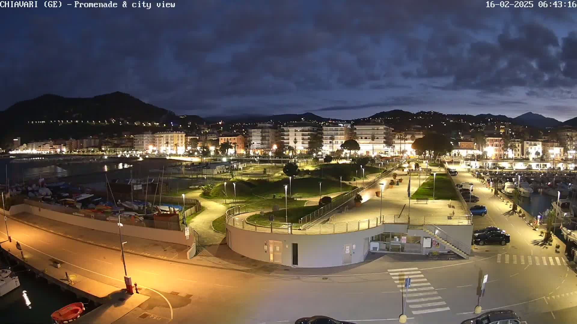 A nighttime, illuminated view of a waterfront promenade and city, with a marina filled with boats and cars parked in a lot, under a partly cloudy sky.