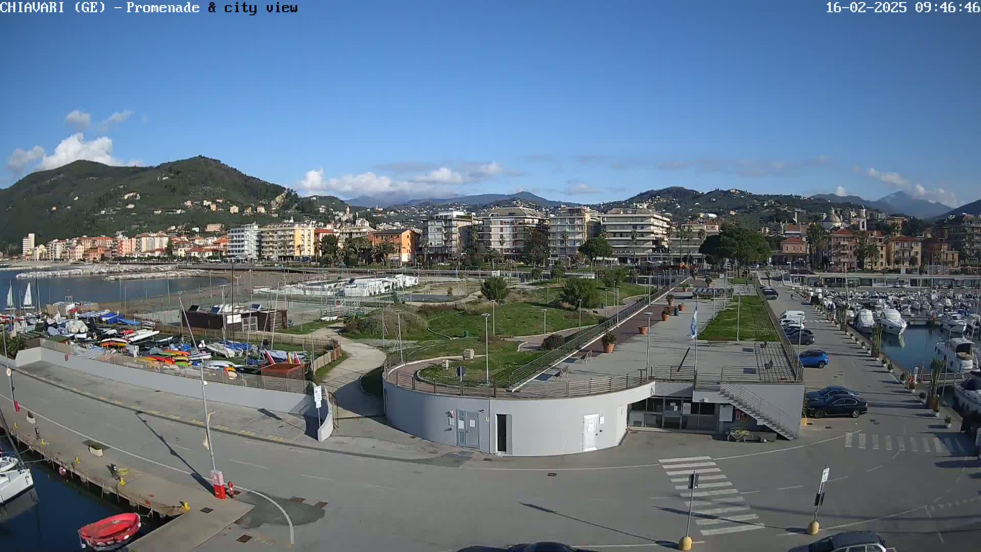 A marina with numerous boats and cars is situated next to a park and a coastal town, all under a clear, sunny sky.