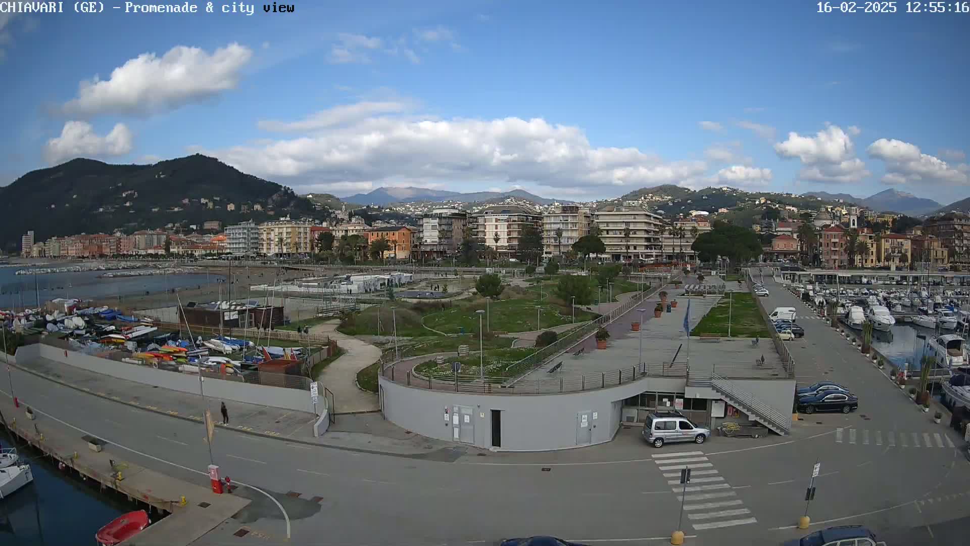 A coastal town on a partly cloudy day, viewed from above, shows a promenade, marina filled with boats, and a park next to a residential area, all situated against a backdrop of mountains.