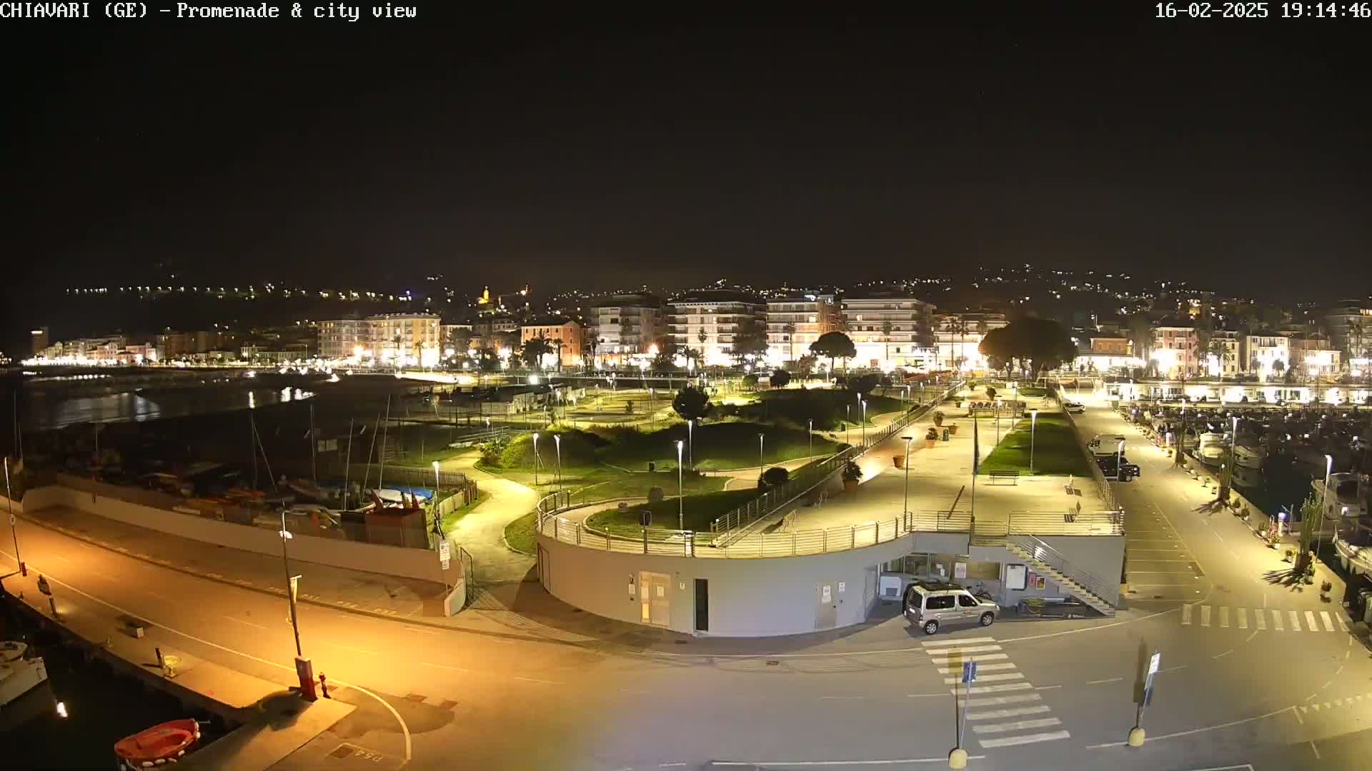 A nighttime view of a coastal city's promenade and harbor, featuring a well-lit park and a marina filled with boats.