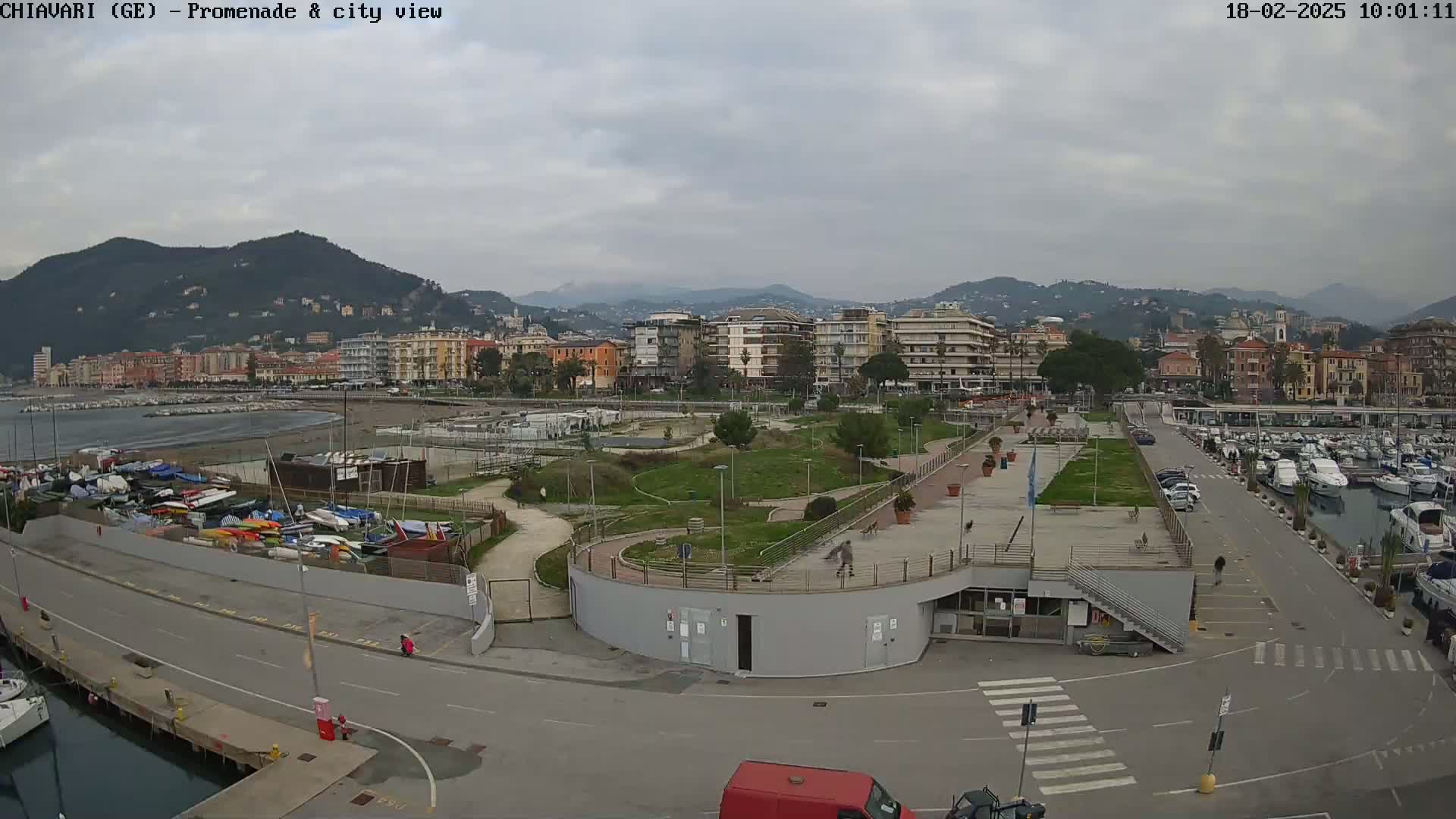 A coastal city scene on an overcast day shows a promenade, park, marina filled with boats, and buildings stretching up a hillside.