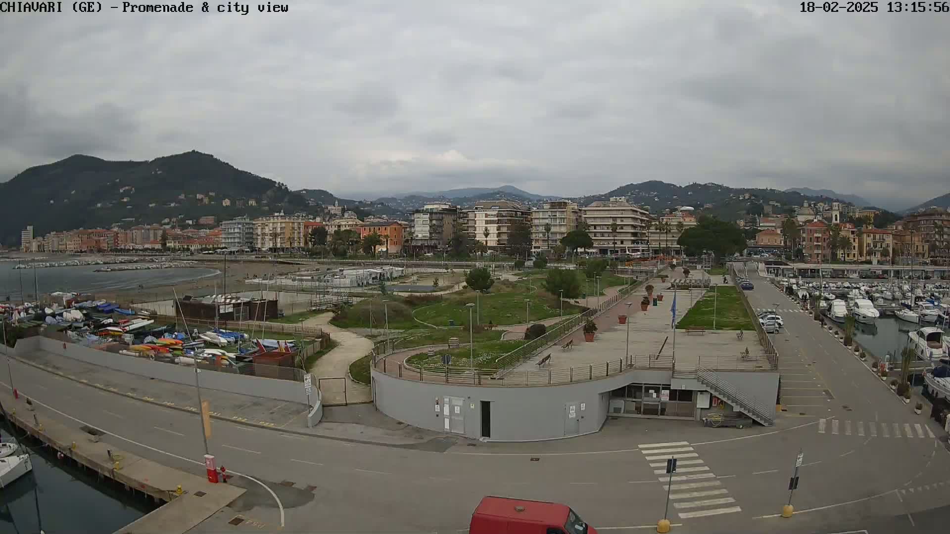 An overcast day reveals a coastal town's promenade, marina filled with boats, and a park-like area, all nestled against a backdrop of mountains.