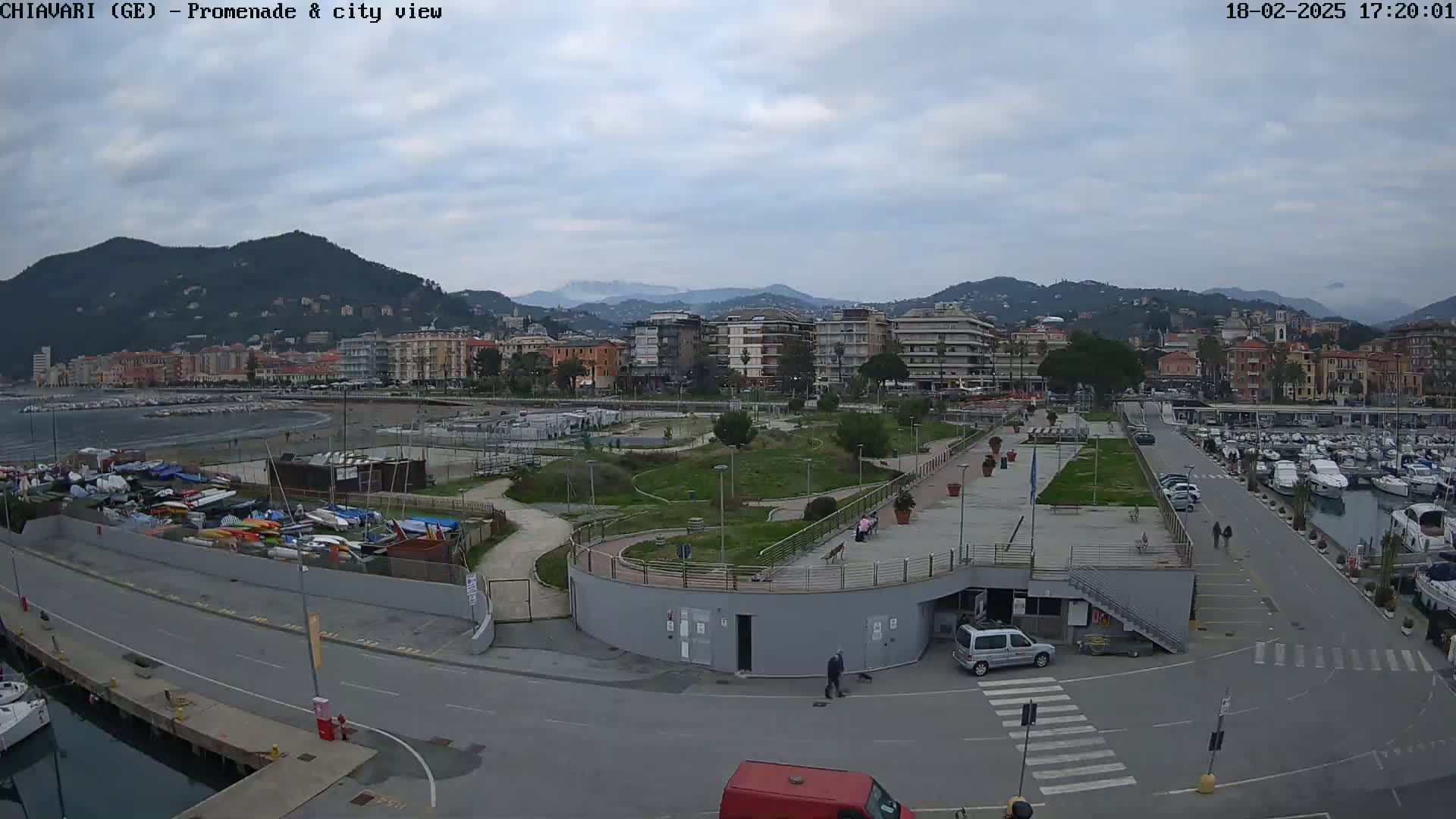 An overcast day reveals a coastal city scene with a marina filled with boats, a promenade, a park, and apartment buildings against a backdrop of mountains.