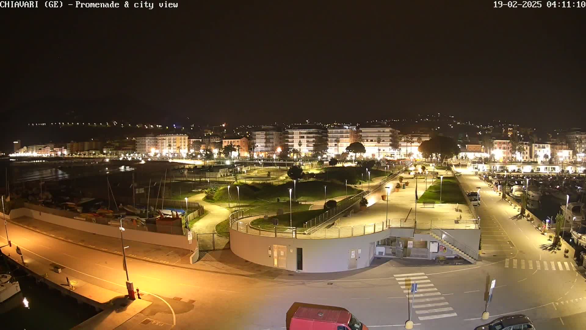 A nighttime, brightly lit view of a waterfront promenade and city, featuring a marina with boats and a park-like area with winding pathways.