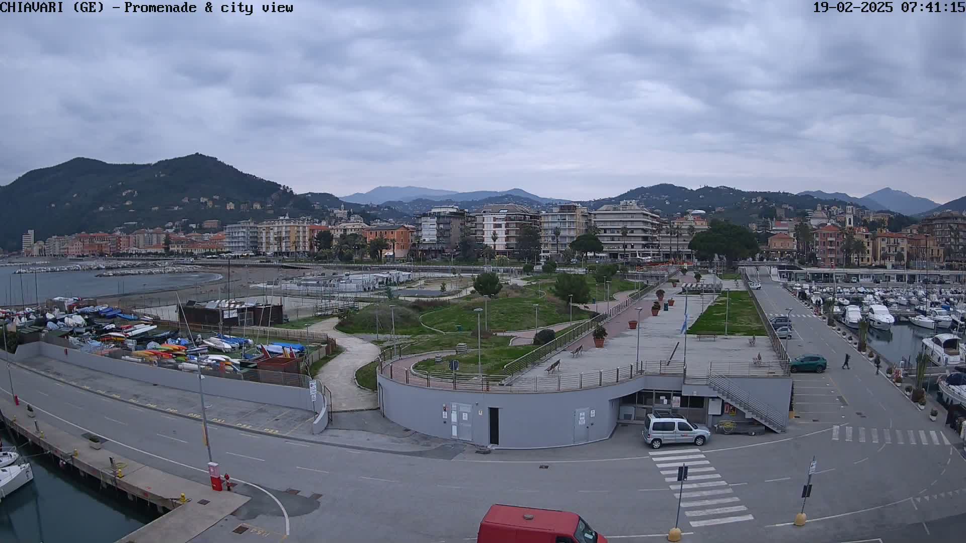 An overcast day reveals a coastal city scene with a marina, promenade, and parkland nestled between buildings and mountains.