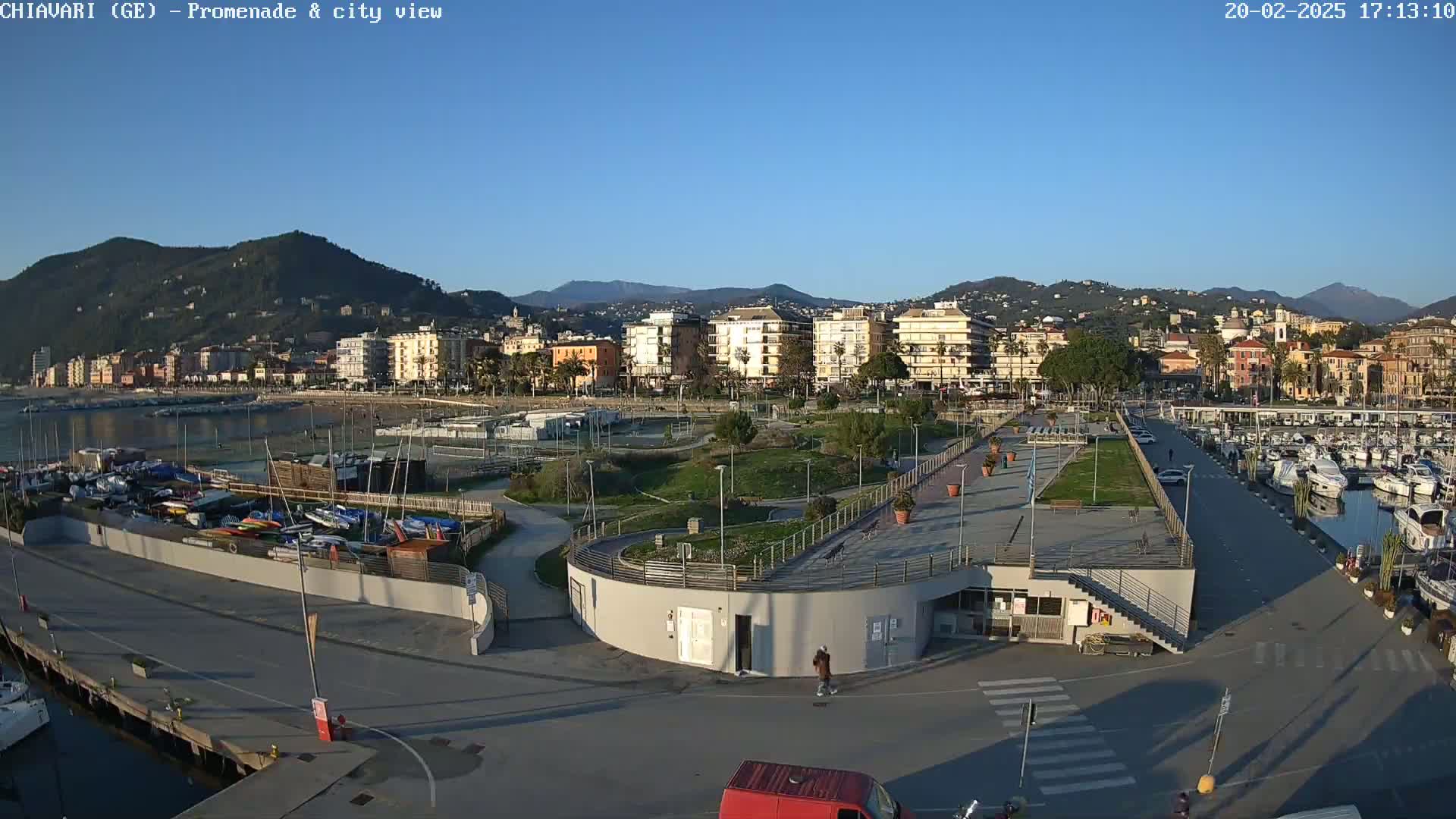 A coastal city scene on a sunny day shows a harbor filled with boats, a promenade with landscaping, and buildings against a backdrop of mountains.