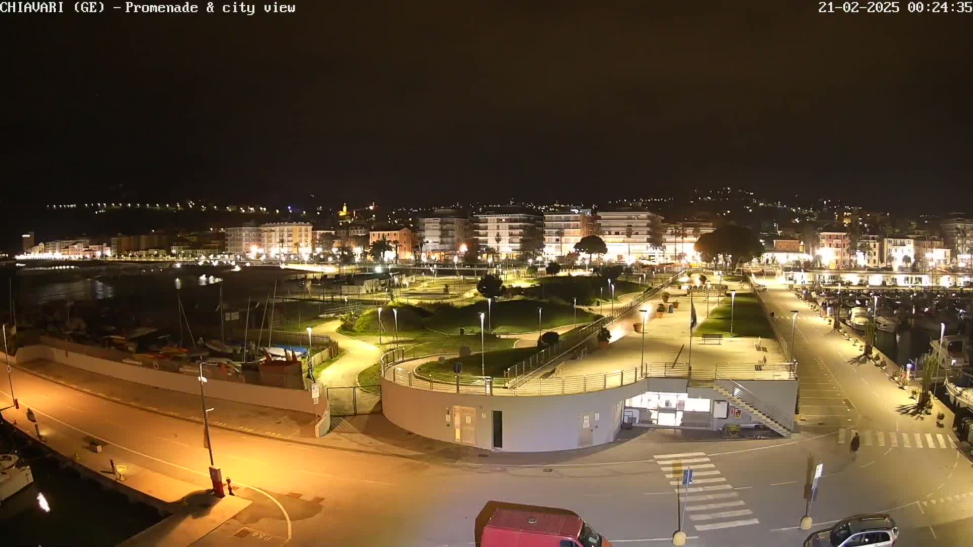 A nighttime, illuminated view of a waterfront promenade and city, featuring a harbor with boats, and a park-like area with winding paths.