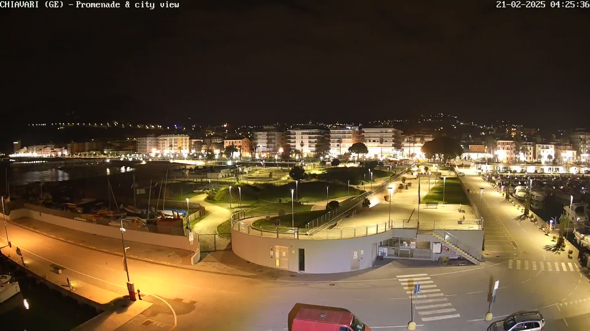 A nighttime view of a city's waterfront promenade and harbor, featuring a landscaped park, parked cars, and boats in the water.