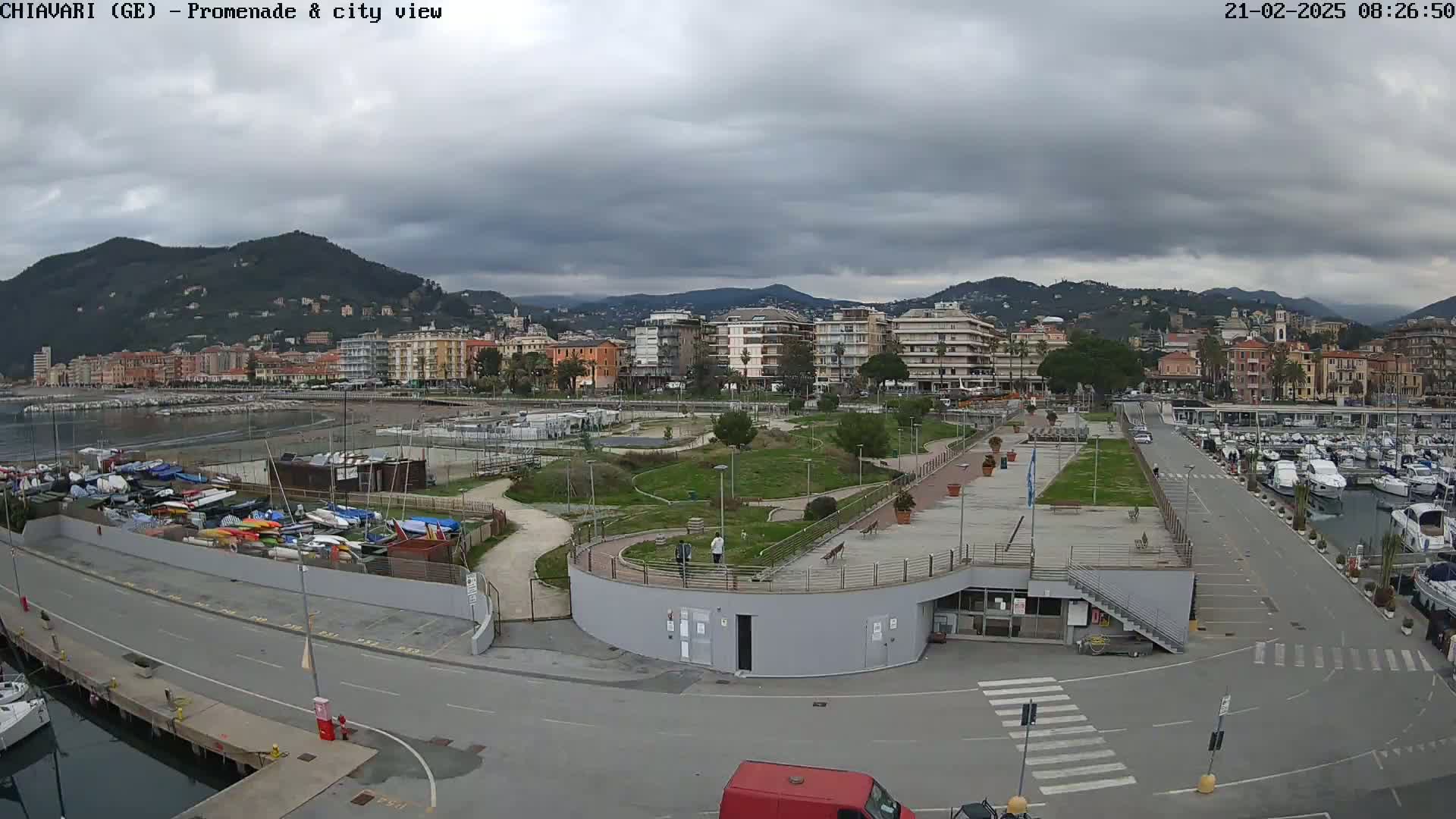 A coastal city scene under a cloudy sky shows a harbor filled with boats, a promenade with grassy areas, and buildings on a hillside in the background.